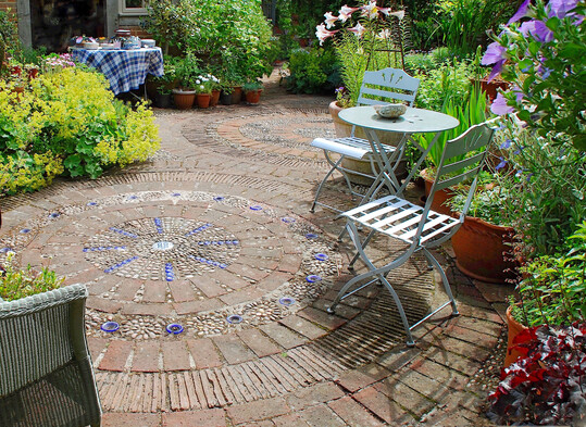 Das Ornament aus Ziegelsteinen, Ziegelriemchen, kobaltblauen Glasringen sowie schmalen und breiten Kieselsteinen ziert den Boden in einem englischen Garten.